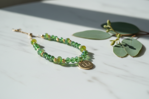 Collana Emerald Glow con cristalli sfaccettati verdi, dettagli oro e ciondolo logo dorato fotografata su una superficie in marmo con foglie di eucalipto – My Pets and I Milano
