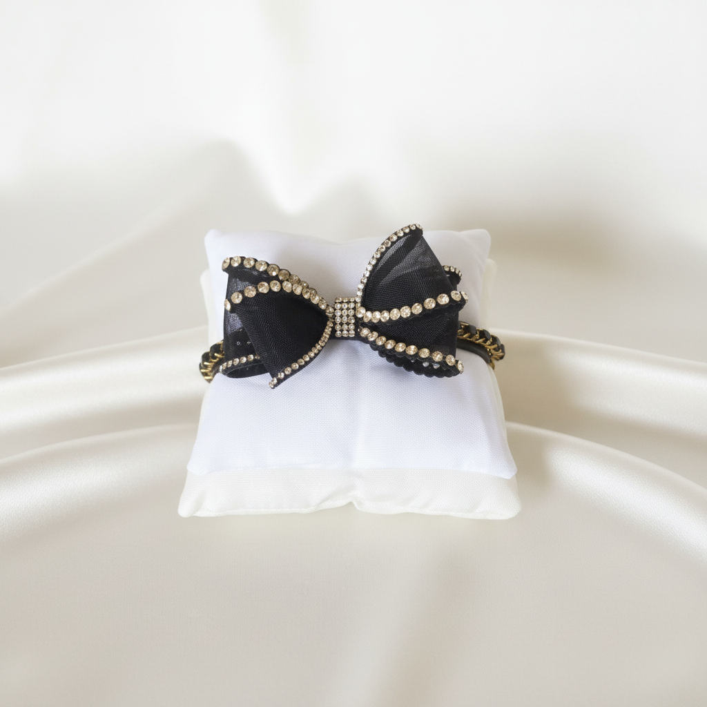 Collare gioiello nero con fiocco decorato da strass e dettagli dorati, disposto su cuscino e sfondo bianco. 
