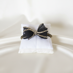 Collare gioiello nero con fiocco decorato da strass e dettagli dorati, disposto su cuscino e sfondo bianco. 