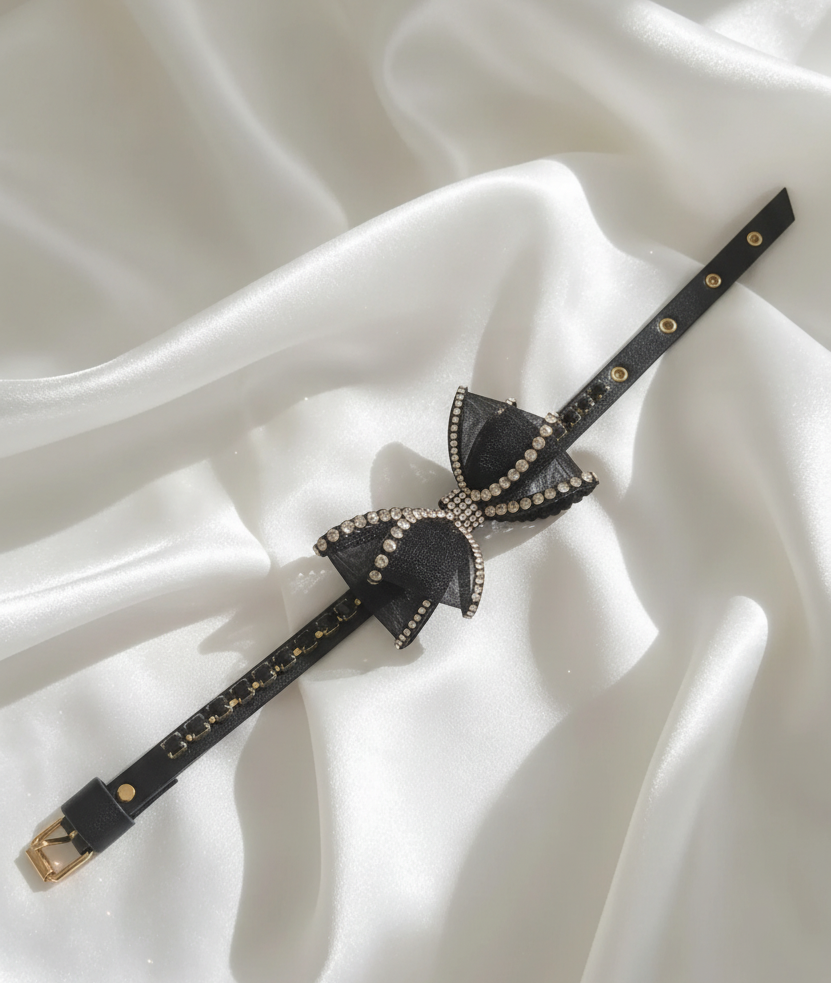 Collare gioiello nero con fiocco decorato da strass e dettagli dorati, disposto su tessuto in seta bianco. 