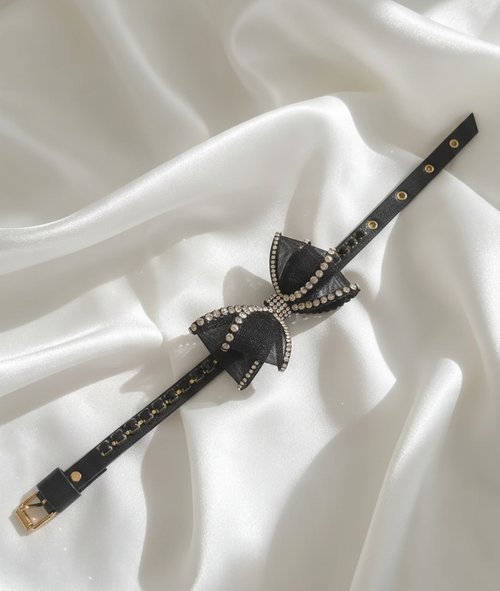 Collare gioiello nero con fiocco decorato da strass e dettagli dorati, disposto su tessuto in seta bianco. 