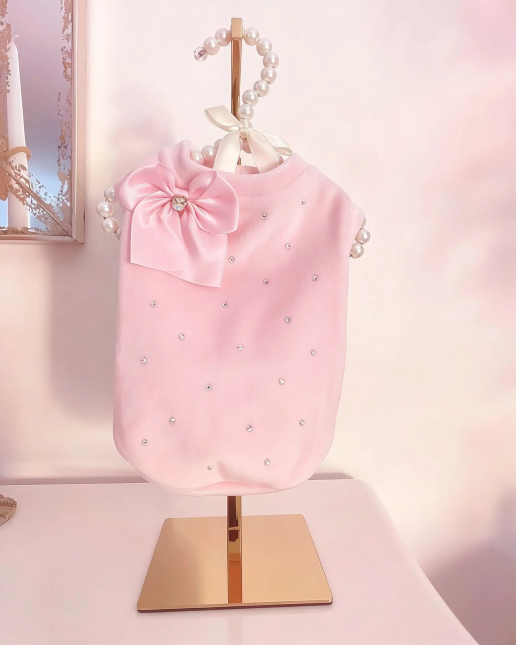 Felpa in ciniglia rosa baby per cani con strass AB e fiocco in raso removibile, esposta su supporto dorato con gruccia decorata da perle.